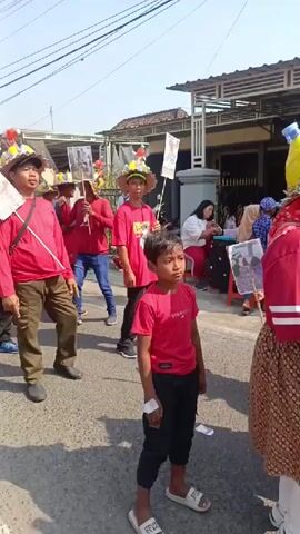 The video shows a street parade. Several people are walking in a procession. They are wearing red shirts and some have on hats decorated with colorful elements. One person in the foreground is a young boy in a red shirt and black pants. Others are carrying flags and posters. In the background, there are houses and people watching the parade from the side of the street. The weather appears to be sunny.