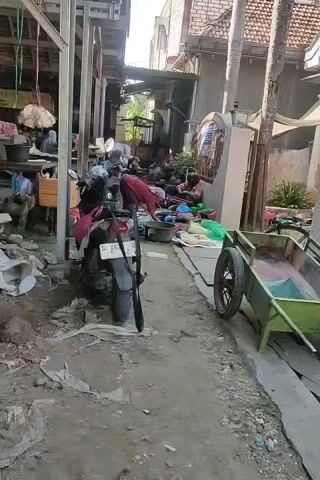The video shows an outdoor market area with several people and motorcycles. The ground is uneven and covered with dirt, debris, and some plastic sheeting. In the foreground, a motorcycle is parked, with a rifle leaning against it. Further into the market, there are stalls with various goods, and people are seen interacting with them. One stall has what appears to be produce or dried goods displayed. There is also a green cart with a wheel visible on the right side of the frame. The overall impression is a busy, somewhat disorganized outdoor market.