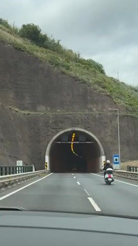 A car is approaching a tunnel in the daytime. A scooter is in front of the car, heading into the tunnel as well. The tunnel is lined with yellow lights and has a concrete interior. Green arrow signs hang over the entrance. The exterior of the tunnel is rocky and surrounded by greenery.