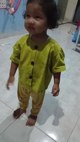 A young child with dark, curly hair tied up in a bun stands barefoot on a tiled floor. The child wears a light green, short-sleeved shirt with three dark buttons down the front and yellow pants. The child's left hand is in their pocket. The background shows a light blue wall with a colorful chart hanging on it, and to the right, a white cabinet with some toys. The lighting suggests an indoor setting.