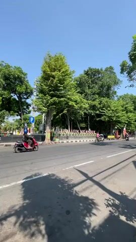 A static shot looking across a street towards a park entrance on a clear, sunny day. The street in the foreground has dappled shade from trees. In the middle ground, there is a sidewalk with a low wall and a large sign that reads "ALUN ALUN NGANJUK" in large white letters. Behind the sign, there are several large green trees. To the left of the sign, there is a blue parking sign. Several motorcycles are visible on the street, with two riders in the middle ground and one in the background. A person wearing a pink shirt is visible behind the sign. The sky is a clear blue.