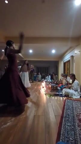 The video captures an indoor gathering in Dharamkot, India, featuring dancing and live music. The scene is set in a spacious room with a wooden floor, plain walls, and recessed ceiling lights. The lighting is dim, enhanced by a line of candles placed along the floor, creating a warm and intimate atmosphere.

In the foreground, several individuals are actively dancing. A woman in a long, dark red dress is seen moving expressively, as is another woman wearing a dark, strapless top and a flowing teal skirt. Their movements suggest a freeform or ecstatic dance style.

In the background, a group of people are seated on rugs on the floor. Some appear to be musicians, with visible instruments including a drum and a stringed instrument that resembles a sitar or similar Indian classical instrument. The seated individuals are observing or participating in the musical performance.

The event appears to be an evening or night gathering, given the reliance on artificial light and candles. The overall mood seems engaging and joyful, with participants immersed in the music and dance. There is no visible text in the video.