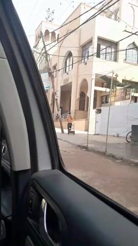 The video, taken from the inside of a car, shows people walking on a street in front of a tan-colored building. The building appears to be a mosque with Arabic writing on its facade. A man wearing a black shirt and jeans walks across the street in front of the car. Another person in a black abaya walks in the same direction. A table with goods for sale is set up on the sidewalk. Electrical poles and wires are visible overhead.