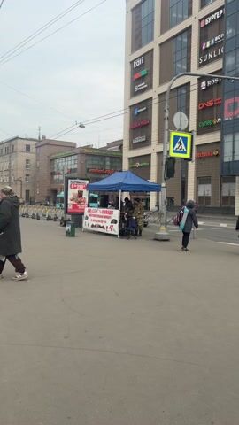 Here is a concise description of the video:

The video shows a group of three men setting up a small performance space under a blue canopy on a sidewalk in what appears to be a city. The men are dressed in camouflage clothing. A sign on the front of their setup indicates that they are a musical group called "ВИА "САЛАНГА" and are performing a charity concert. A telephone number is displayed on the sign. Next to the canopy is an advertisement for a fast-food combo meal and a green recycling bin with a poster on it. In the background, there are several buildings with storefronts and signs in Russian. Traffic is moving in the street nearby.