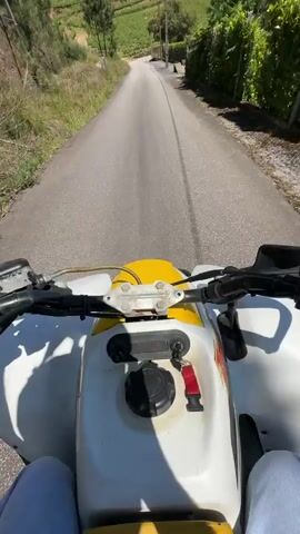 The video shows a first-person perspective of someone riding a white and yellow ATV down an empty asphalt road. The road leads through a rural area with terraced fields and scattered houses. Green vegetation, trees, and a wire fence border the road.  The sky is a clear, bright blue.  The rider wears blue jeans, and their hands are visible on the handlebars of the ATV. The vehicle is in motion.