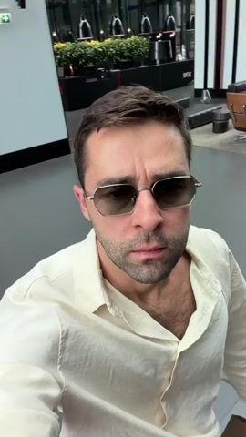 The video shows a man wearing sunglasses and a cream-colored shirt. He is indoors, possibly in a shopping mall or airport, as indicated by signs and displays in the background. The man is filming himself in a selfie-style video, with the camera moving slightly as he turns his head and smiles. The lighting is bright and natural. The background is blurred, suggesting a shallow depth of field. There are no other people visible in the frame.