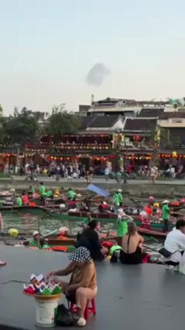 The video shows a bustling waterfront scene, likely a tourist destination, with several boats on the water. Many of the boat operators are wearing green shirts and orange life vests. People are strolling along the waterfront and some are sitting on the edge. In the foreground, a woman wearing a polka-dot hat is selling small, colorful items from a basket. Behind the waterfront, there are buildings adorned with lights. The sky is a light color, suggesting it is either early morning or late afternoon.