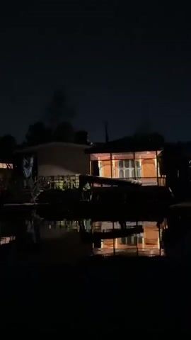 This is a dark, wide shot of a houseboat at night. The houseboat is illuminated from within by warm, orange lights, and its reflection is visible on the still, dark water in front of it. Silhouetted trees are visible behind the houseboat against the night sky. The scene is static and the lighting suggests it is late at night.