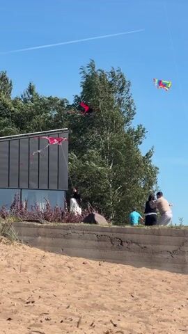 A group of people are on a sandy beach flying kites under a clear blue sky. A modern-looking building with a dark, textured wall is visible on the left. Green trees are behind the building and the people. There are at least three kites in the air. One is red and black and appears to be a stunt kite. Another is a colorful delta kite. A third kite, also colorful, is further away and higher in the sky. Several people are standing and sitting on a concrete ledge overlooking the beach. One person, dressed in white, is holding a kite string and appears to be launching or flying a kite. Another person in a blue shirt is sitting on the ledge. A couple is sitting together on the ledge, with one person wearing a white top and the other in dark clothing. The scene is bright and appears to be a sunny day.