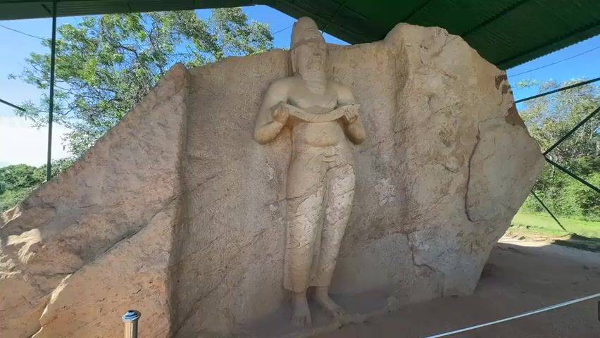 The video presents a medium shot of an ancient rock carving sheltered under a green metal roof. The carving, a human figure, is integrated into the natural rock face. The figure is male, appears to be holding a scroll or tablet, and wears what appears to be a simple, draped garment. The rock surface is rough and textured, with variations in color. Beyond the roof structure, glimpses of trees and sky suggest an outdoor setting. A bollard is seen on the ground near the rock formation.