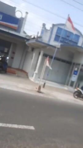 A slightly blurred, low-resolution video captures a street scene, possibly in Indonesia given the presence of Indonesian flags. The video appears to be shot from a moving vehicle. In the foreground, a paved road with a white line is visible. In the middle ground, a row of buildings lines the street. The first building on the left is identifiable as a BRI bank with its distinctive blue and red logo. Several motorcycles are parked in front of the buildings, and a few people are seen near the bank entrance and by the side of the road. Further down the street, more buildings and parked motorcycles are visible. Red and white Indonesian flags are hung along the street, suggesting a celebratory occasion. The sky is overcast.