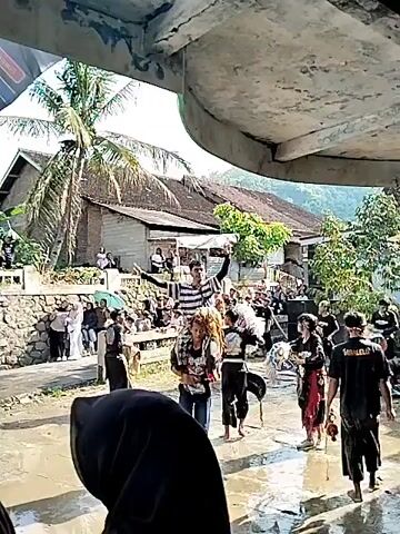 The video shows a cultural performance taking place outdoors. The ground appears to be wet, possibly from rain, and people are gathered around to watch. Performers in dark clothing and masks are moving around, with one man being carried on the shoulders of another. A crowd of people are observing from behind a low stone wall, and the performance is happening in a village-like setting with houses and trees visible in the background. There’s a banner visible in the scene, and the video is taken from under a structure, possibly a roof or awning.