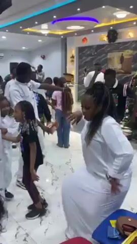 The video shows a group of people dancing in what appears to be a church or event hall. Many individuals are wearing white attire, while others are dressed in casual clothes. The room is brightly lit, with modern lighting fixtures in the ceiling and decorations on the walls. People of various ages are present, including young children who are also dancing. There is music playing in the background, and the atmosphere seems lively and celebratory. Some people are holding phones, possibly recording or taking pictures. There are chairs arranged around the room, and some objects such as purses or bags placed on them.