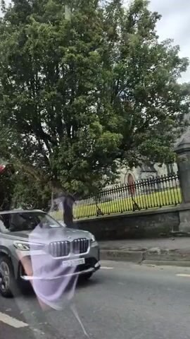 This media file captures a scenic view from a moving vehicle in Innishannon, Ireland. The primary focus is on a church with a tall, pointed spire, characteristic of Gothic architecture. The church is constructed of stone and appears to be quite old, with arched windows and a wooden door visible. It is surrounded by greenery, including trees and grass, and is separated from the road by a decorative black metal fence and a stone wall.

Several vehicles are visible, including a light-colored SUV, a dark grey hatchback, and a black Audi. A white sheet or fabric is draped over the rear wheel of one of the vehicles, suggesting a potential cover for storage or protection. The sky is cloudy, indicating overcast weather. The overall atmosphere is calm and serene, typical of a drive through a rural or semi-rural area. There is no visible text in the media file.