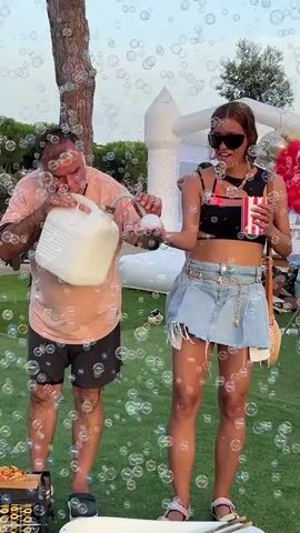 A man in a pink t-shirt and dark shorts is blowing bubbles. He is holding a white plastic jug with a handle in his left hand and a white circular object in his right hand. He is blowing air into the circular object, creating bubbles. He is standing on a green grassy lawn. To his right is a woman wearing a black crop top, a denim skirt, and sunglasses. She is holding a red and white striped cup in her right hand and a popcorn container in her left hand. She is smiling. In the background, there is a white inflatable bounce house, some trees, and people sitting on chairs. There are many bubbles floating in the air around the man and woman.