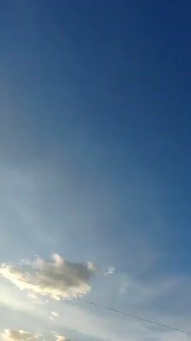 The video shows a person holding a phone vertically and recording the sky. The sky is blue with some scattered white clouds. There are some thin, wispy clouds in the distance. In the lower part of the frame, there are some power lines and silhouetted trees and buildings. The sun is setting, casting a warm glow on the clouds and trees. The person's voice can be heard saying, "The sky is so cloudy, so cloudy."