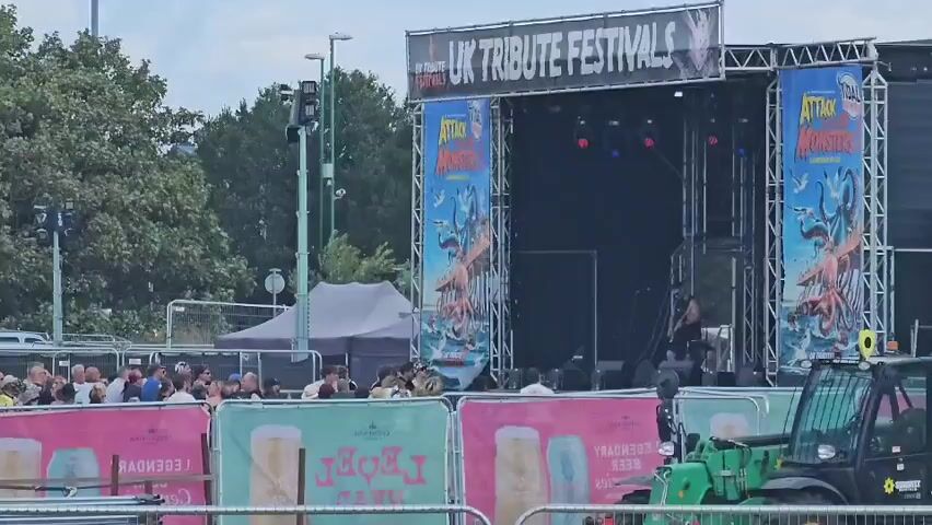 The video shows an outdoor music festival stage with a large banner overhead that reads "UK TRIBUTE FESTIVALS". Two tall banners on either side of the stage depict a monstrous creature rising from the sea, with the text "ATTACK OF THE MONSTERS" above it. A band member is visible on stage, seemingly preparing to play. In the foreground, there are crowd barriers with advertisements for "LEGENDARY BEER" printed on them. A few people are gathered behind the barriers, watching the stage. A green forklift is parked to the right of the frame. The environment appears to be a festival grounds, with trees and some temporary structures visible in the background. The sky is overcast.