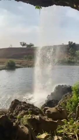 A waterfall cascading into a pond is viewed from a rocky overhang. The water falls into the pond in a single stream, creating a splash at the bottom. The pond is surrounded by vegetation and a brown field with a few trees in the background. The sky is clear and bright.
