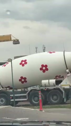The video shows a white cement mixer truck with red flower decorations parked on the side of a road. The truck has the logo "Marera CONSTRUCTION" on it. In the background, there is a construction site with a crane and some buildings. A black car passes by on the right side of the frame. The weather appears to be cloudy. The video is shot from inside a car.