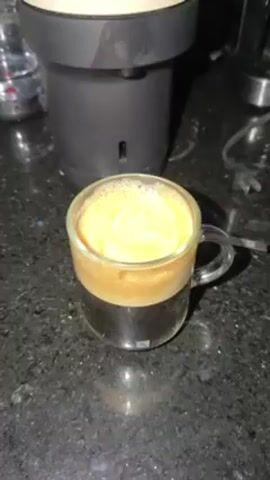 A close-up shot shows a clear glass mug filled with coffee and a creamy, light-brown foam on top, sitting on a black countertop with speckled patterns. Behind the mug is a Nespresso coffee machine in cream and dark-gray colors, as well as a bottle of water. A stainless-steel pitcher with the word "NESPRESSO" printed on it is shown pouring frothy white milk into the coffee mug. The countertop appears clean and well-lit.