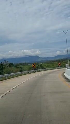 The video shows a view from a vehicle traveling on a curved asphalt road. The road has white and yellow lane markings. There are guardrails on the sides of the road. In the distance, there are green hills and mountains under a cloudy sky. There are also several streetlights visible. A yellow curve warning sign is present on the left side of the road.