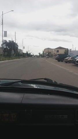 The video, filmed from inside a moving vehicle, shows a view of a road and its surroundings on a cloudy day in Borodulikha Rural District, Kazakhstan. The perspective is through the windshield, with part of the dashboard and a windshield wiper visible on the right side of the frame. The word "LADA" is discernible on the vehicle's interior.

The vehicle is traveling down a paved, two-lane road that appears to be a main thoroughfare. On the left side of the road, there are several buildings, including a prominent light-colored structure with a brown roof that shows signs of age. Further down the street, more buildings and trees line the roadside. Streetlights are also visible at intervals.

Traffic on the road is light. At the 0:02 mark, a white car with its headlights on approaches and passes the filming vehicle. Another dark-colored car with headlights illuminated is seen approaching around the 0:04-0:05 mark. The sky is heavily overcast with significant cloud cover, indicating a dull daytime. The overall scene suggests a small town or rural settlement.
