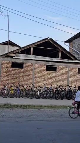 A row of bicycles is parked outside a brick building with a wooden roof. To the right, a modern building with large glass windows stands adjacent to the brick structure. A young child is visible near the entrance of the modern building, interacting with some of the parked bicycles. The scene appears to be outdoors with a clear sky overhead and a paved road in the foreground.
