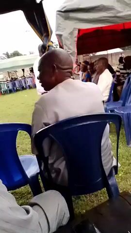 A group of people are seated under a tent on a grassy area. The tent is made of white fabric with red trim. The people are mostly men, and some are wearing traditional African clothing. Some are looking towards the left of the frame, where a man in a light-colored shirt appears to be speaking. The overall mood seems to be one of a gathering or event.