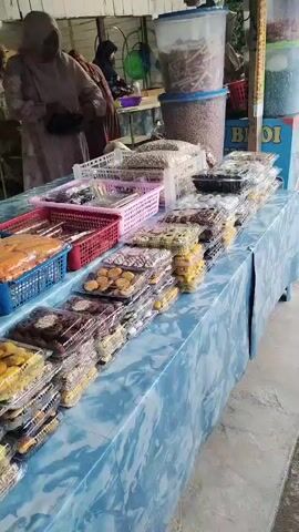 The video shows a table covered in a light blue marble-patterned tablecloth, laden with various types of cookies and snacks packaged in clear plastic containers and baskets. The cookies are of different colors and designs, neatly stacked. In the background, there are two women wearing hijabs, presumably vendors, interacting with customers. Behind them, shelves hold larger containers of goods. The setting appears to be a market stall or a temporary outdoor sales point.