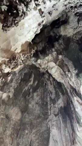 The camera pans down from the ceiling of a cave to reveal a brightly colored temple. The cave ceiling is mottled and textured with a mix of gray and white tones. A sign reading "PAAL KUDAM WAY" in English and another language hangs above the temple entrance. The temple is decorated with multiple tiers, each brightly lit with colorful lights. Several people can be seen entering and exiting the temple grounds. A sign indicating "NO SHOES" is visible near the entrance. A security guard wearing a yellow vest stands nearby. The temple appears to be located inside the cave.