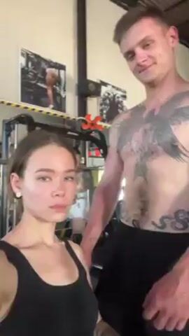 Here's a description of the video:
A young woman and a shirtless man stand close together in a gym. The woman, wearing a black tank top, flexes her arm to show her bicep, while the man, who is taller and has several large tattoos on his chest and abdomen, flexes his bicep as well. They look at each other and smile. The background includes gym equipment and posters.
