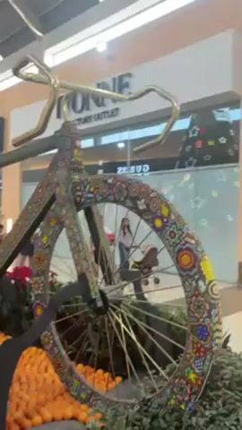 The video shows a large bicycle decorated with colorful patterns that are reminiscent of Huichol artwork. The bicycle appears to be made of metal and the frame, wheels, and other parts are covered in vibrant designs. It is displayed indoors in a shopping mall. In the background, pedestrians walk by storefronts such as "IVONNE FACTORY OUTLET". The base of the bicycle display is surrounded by small orange pumpkins and red flowers. The lighting is bright and artificial.