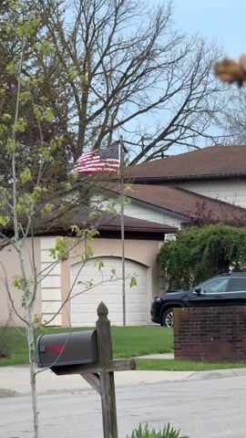 The camera captures a view of a suburban street scene in daylight. An American flag flies atop a pole in the background, partially obscured by bare tree branches and a nearby house. In the foreground is a weathered wooden mailbox post with a dark-colored mailbox attached. The garage of the house behind the flag is visible, as is a parked black SUV. Fragments of speech can be heard in the audio, indicating the presence of people, although they are not visible in the frame.