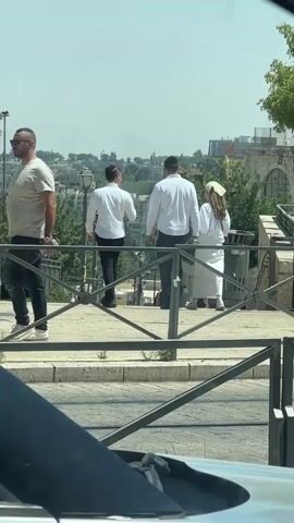 The video shows a bright, sunny day in Jerusalem, Israel, with a clear, hazy sky. Three individuals are walking away from the camera on a paved pathway lined with a metal railing. The two individuals in front are men wearing white shirts and dark pants. The person behind them is a woman wearing white and a light-colored head covering.

In the background, there is a cityscape with buildings and trees, suggesting an urban environment. The architecture appears consistent with Jerusalem, featuring stone buildings and a hilly landscape. A lamppost is visible on the left side of the pathway. Further in the background, more buildings and greenery are spread across rolling hills.

As the video progresses, other people become visible walking on the path. Two men, dressed in casual attire, are seen interacting. One is wearing a light-colored polo shirt and light pants, while the other is wearing a dark t-shirt. Another person, dressed in dark clothing, walks by on the right.

The overall scene depicts a leisurely stroll through a scenic area of Jerusalem on a warm, sunny day. The light suggests it is mid-day. There is no visible text in the video.