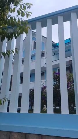 The video pans across a pale blue and white modern house with a slanted roof. The house is partially visible through a white fence with wide vertical slats, topped with a pale blue rail. Green foliage and purple flowering bushes are visible in the yard behind the fence. The sky is overcast.