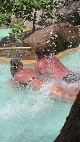 A couple is filmed from behind while partially submerged in a pool of light turquoise water. They are both face-down and appear to be swimming, with the man slightly ahead of the woman. The woman is wearing a black and white bikini, and the man is wearing dark swim trunks. Water droplets are spraying in the foreground, creating a blurry effect. Behind them, there's a decorative rock structure, along with some green foliage. The sunlight is bright, highlighting their skin. The scene is recreational and appears to be taking place at a water park or resort.