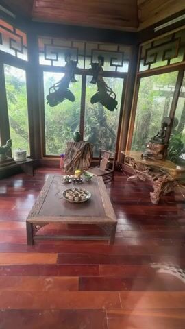 The video shows the interior and the view outside a room with large windows that offer a view of lush greenery and a small town. The room has a wooden floor and is furnished with rustic wooden furniture, including a low table, a chair, and decorative pieces. A set of teacups is arranged on the table. The video then pans outside, revealing a view of a landscape with trees and a small town in the distance. The scene appears peaceful and tranquil.