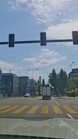 A first-person perspective video shows a daytime drive through a city. The sky is blue with scattered white clouds. On the left side of the road are modern buildings, including one with an "Ibis" sign. On the right, tall green trees line the street. A white van drives on the road ahead, followed by a white truck with a closed trailer. The car passes yellow crosswalk markings. Streetlights are visible along the road.