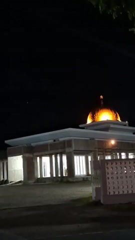 The video shows an exterior shot of a mosque at night. The mosque has a prominent golden dome at its center, which is brightly lit from within or below, making it glow orange-yellow against the dark sky. The main structure of the mosque appears to be in an incomplete state of construction, with visible concrete or unfinished walls, though the interior is illuminated with white light. A white fence with decorative cutouts is visible in the foreground to the right, separating the mosque grounds from what appears to be a paved road. The sky is dark, with a few faint light sources visible, possibly stars or distant lights. A few tree branches are visible at the top of the frame. The overall scene is static, with no people or significant movement. Sounds of a call to prayer (Azaan) are audible.