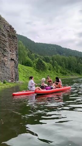 A video shows three young women and a white dog on two red paddleboards floating on a wide river. The women are seated and appear to be chatting and enjoying the scenery. One woman is holding a paddle, while another appears to be taking a photo. The dog is sitting calmly between two of the women. The river is surrounded by lush green trees and a rocky cliff face on the left side of the frame. The sky is overcast with dramatic clouds. The water is dark and reflects the surrounding landscape. The overall atmosphere of the video is relaxed and serene, capturing a moment of leisure and connection with nature.