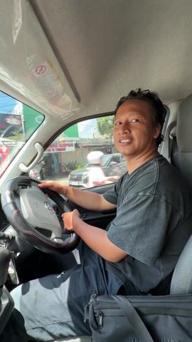 A man is sitting in the driver's seat of a vehicle, holding the steering wheel. He is wearing a dark gray t-shirt and dark pants. His hair is pulled back, and sunglasses are on his head. He looks over his left shoulder, smiles, then looks forward. The car appears to be parked on a street with buildings and other vehicles visible through the windows. A small black bag is visible on the passenger seat.