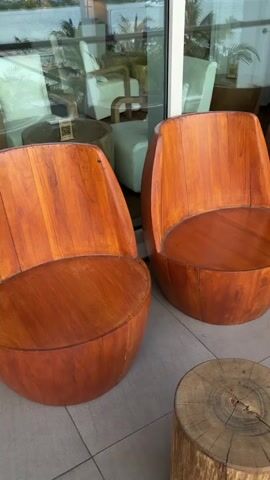 The video pans across an outdoor seating area with a tiled floor. There are several wooden chairs with curved backs and round seats, resembling hollowed-out logs. Beside the chairs, there are cylindrical wooden stools of varying heights, also resembling tree stumps. In the background, a wooden fence with vertical slats separates the seating area from what appears to be an indoor space with large windows reflecting the outdoor environment. Potted plants are visible in the background on the right side of the frame. No people are present. The lighting suggests it is daytime.