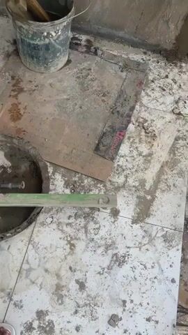 This video provides a brief tour of a property under construction in Angwan Sayawa, Nigeria.

The initial frames (0-7 seconds) show the interior of the building, featuring newly laid white and gray marble-patterned floor tiles that are soiled with construction debris. Various tools and materials are visible, including a circular tub containing cement mix and a trowel, a spirit level, tangled metal wires, and a bucket filled with wooden scraps. A person's bare foot is partially visible in the bottom left corner at the beginning of the video. The walls throughout these interior shots are rough and unpainted, indicating ongoing construction or renovation. A black-framed doorway leads into an adjacent room where the tiled flooring continues, reflecting light.

The final shot (7-9 seconds) transitions to an exterior view, seemingly from a balcony or an open section of the building. A concrete balustrade is in the foreground, and the same bucket with wooden pieces is present. The background reveals a panoramic view of the surrounding area, featuring other buildings (some appearing to be under construction), green landscape, and distant hills or mountains under a cloudy sky.

The video depicts an active construction site, showcasing the unfinished state of the building's interior and its external environment. The time of day appears to be daytime, under natural light. No specific activity beyond the visual sweep of the site is shown, and no text is visible.