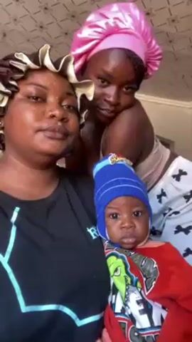 The video shows three individuals: two women and a baby. One woman, wearing a black shirt with a design and a brown hair bonnet, is holding the baby, who is dressed in a red shirt and a blue hat. Another woman is behind her, wearing a pink hair bonnet, a beige top, and white and black patterned pants. The woman in front looks up initially, then looks directly at the camera. The woman behind her maintains a steady gaze at the camera. The baby looks directly ahead with a serious expression. The background includes a patterned ceiling. The woman in front raises her hand and makes a waving motion toward the camera.