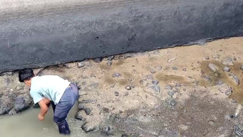 A young boy in a light blue t-shirt and dark pants crouches in muddy water near a concrete wall. He is reaching into the water with his right hand. Rocks and debris are scattered around the muddy area. The water appears murky.