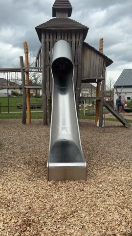 Two boys slide down a tall metal slide on a playground. The older boy, around age 13, wears a white t-shirt, jean shorts, and red slides with white socks. The younger boy, approximately 3 years old, is in the older boy’s arms and wears a green baseball cap, a black jacket with neon yellow accents, denim shorts, and white shoes. They both exit the slide smiling. The older boy puts the younger boy down, and he immediately falls to the ground. The slide is part of a wooden play structure in a playground covered in wood chips. There is a cloudy sky above, and there is a park with benches in the background.