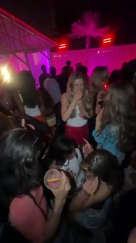 The video depicts a lively night scene at an outdoor club or party, illuminated by vibrant purple lighting. A crowd of young adults, predominantly women, are dancing and socializing. A woman in a white tank top and red skirt is centrally featured, laughing and dancing energetically with her arms raised. Other individuals are seen dancing around her, holding drinks. The scene transitions to two women sitting on a light-colored surface. One woman, with dark hair and wearing a white long-sleeved top, is facing and interacting with another woman wearing a striped dress and holding a drink. The overall environment is energetic with people moving and conversing.