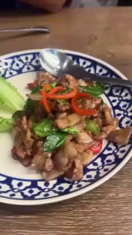 The video shows a spread of dishes on a wooden table in a restaurant setting. A plate with stir-fried pork belly, garnished with green peppers, red chili rings, and lettuce, is visible. A white plate of plain rice sits next to it. Another tray holds small, yellow fried dumplings tied with a string, accompanied by a small bowl of red dipping sauce. A fork and spoon are placed near the plates, indicating that someone is about to eat. The table is simply decorated, and another person's arm is partially visible in the background, suggesting a shared dining experience.