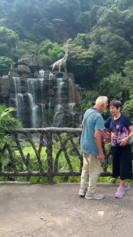 A middle-aged couple is in the foreground, standing on a pathway in front of a wooden railing. The man, wearing a striped blue and white shirt and light-colored pants, has his arm around the woman. The woman, wearing a dark blue top and carrying a bouquet of flowers, is leaning her head towards the man. Behind them, a multi-tiered waterfall cascades down a rocky cliff face. A large dinosaur statue is perched atop the waterfall, its long neck extended. Lush green foliage surrounds the waterfall and dinosaur, creating a naturalistic backdrop. The overall scene appears to be a scenic attraction.