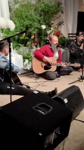 A group of people are performing music indoors. In the foreground, a man with a beard is playing an acoustic guitar and singing. He is wearing a red shirt. To his right, a person is sitting and singing into a microphone. Behind them, two people are playing drums. One is wearing a black hoodie and the other is wearing a green jacket. A keyboard is visible in the background. There are speakers and musical equipment on the floor. The room has a stage-like setup with curtains and decorations. The lighting is warm and intimate.